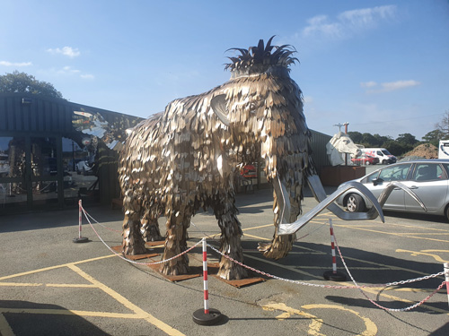 A detailed metal sculpture of a mammoth at the British Ironwork Centre, showcasing its large curved tusks and shaggy appearance, set in the outdoor exhibition area.