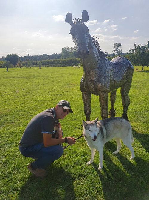 A whimsical metal sculpture of a house at the British Ironwork Centre, featuring figures of 'me' and 'Mia' admiring the artwork, set in the outdoor exhibition area.