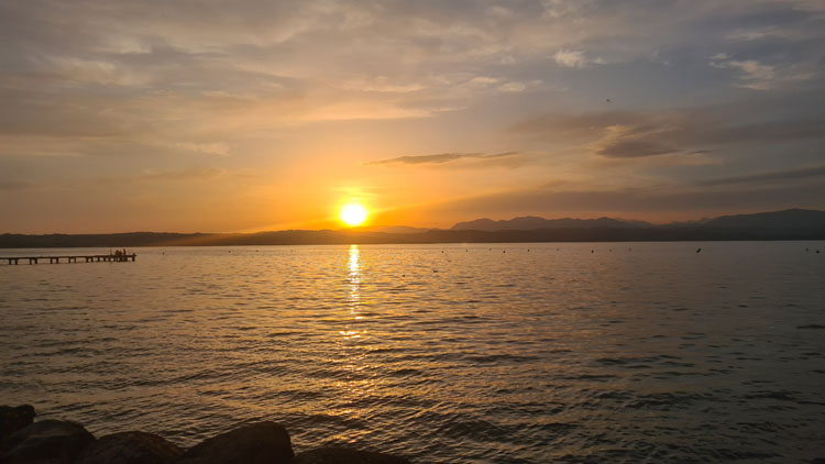Peaceful sunset over a calm lake with hazy mountains, soft sky hues, a short pier, and subtle reflections on rippling water.