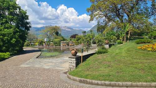 The image depicts a serene scene of a stone path meandering through a lush garden at the Giardini Botanici Di Villa Taranto. Flanking the path are vibrant trees and grass, with a tranquil pond reflecting the sky and clouds above. This picturesque landscape invites visitors to enjoy nature's beauty while offering seating areas like benches throughout the park.