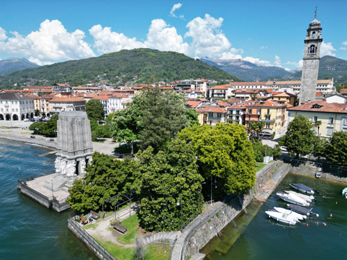 The image captures a scenic view of Lake Maggiore, featuring a body of water with boats gently floating. In the background, various buildings and possibly a lighthouse are visible, adding to the city's charm. The landscape is framed by trees, mountains, and a beautiful sky adorned with clouds.