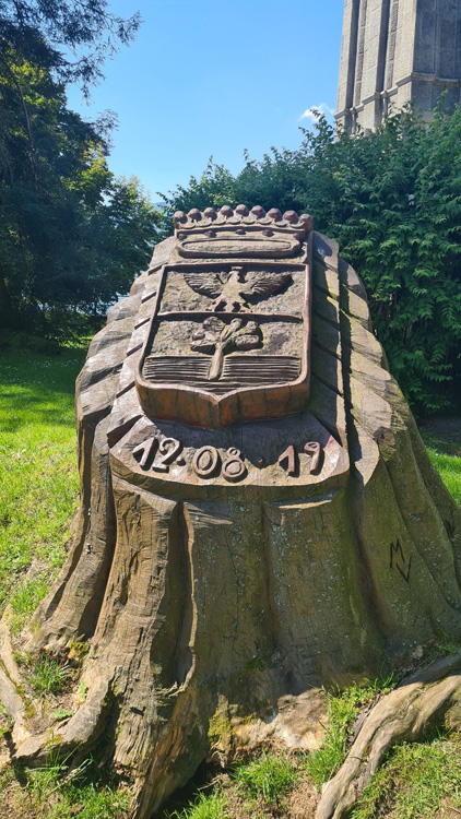 The image features a carved stone displaying a crest and some text. It was captured outdoors at Lake Maggiore, surrounded by elements of nature such as grass, trees, and the sky. The serene setting enhances the historical significance of the statue-like stone. The date provided is December 8th, 2019.