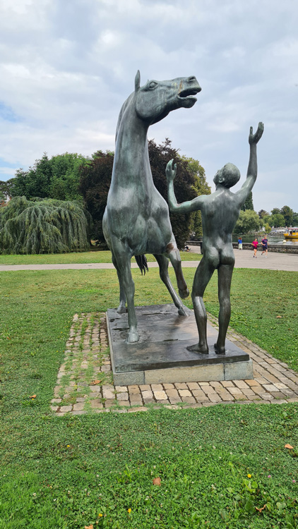 Bronze statue of two horses in a Geneva park, surrounded by grass, trees, and a cloudy sky, with a person standing beside it