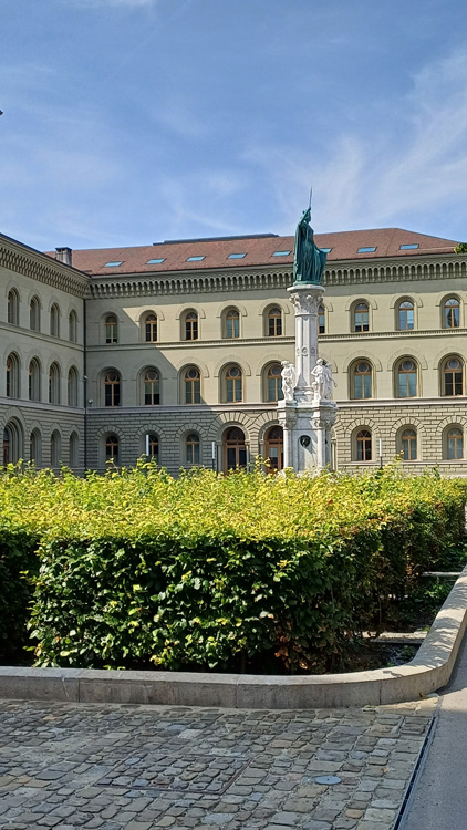 Large building with a prominent tower, stone facade, and surrounding garden area under a blue sky