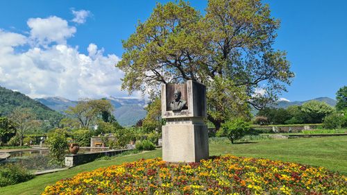 The image depicts a statue of a man situated in a garden. The surrounding environment features outdoor elements such as trees, grass, and flowers, set against a backdrop of sky and clouds. Additionally, the landscape includes mountainous terrain and is part of the Giardini Botanici Di Villa Taranto.