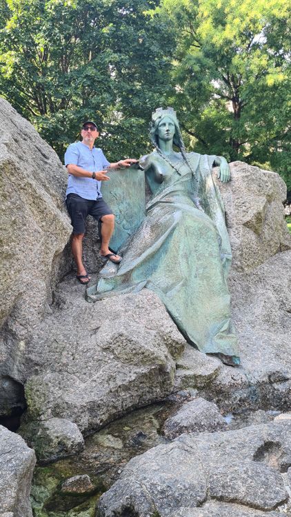 Man posing beside the Universal Post Monument in Bern, surrounded by trees and rocky terrain on a hiking trail