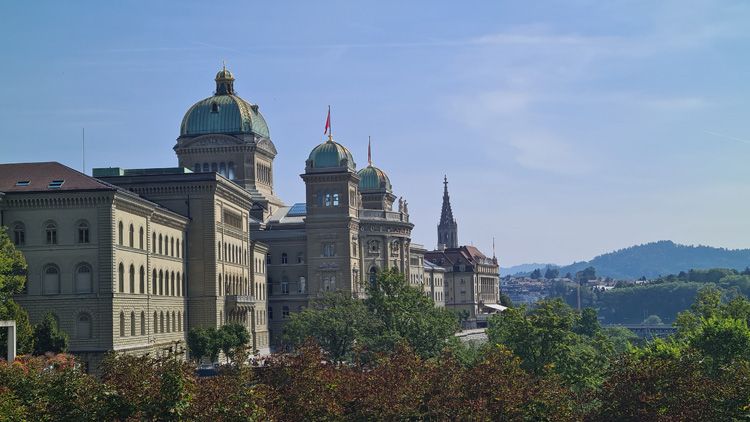 Grand building with a clock tower, domes, and castle-like features, surrounded by trees and mountains under a cloudy sky