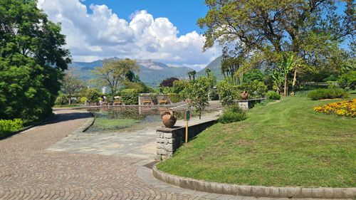 The image depicts a serene scene of a stone path meandering through a lush garden at the Giardini Botanici Di Villa Taranto. Flanking the path are vibrant trees and grass, with a tranquil pond reflecting the sky and clouds above. This picturesque landscape invites visitors to enjoy nature's beauty while offering seating areas like benches throughout the park.