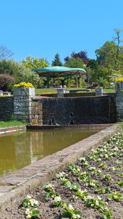 The image features a picturesque pond with a gazebo situated in the background. Surrounding the pond are lush plants and trees, contributing to the outdoor ambiance. The scene captures a serene moment with clear water reflecting the sky, creating a tranquil atmosphere in this beautiful botanical garden setting, likely at Giardini Botanici Di Villa Taranto.