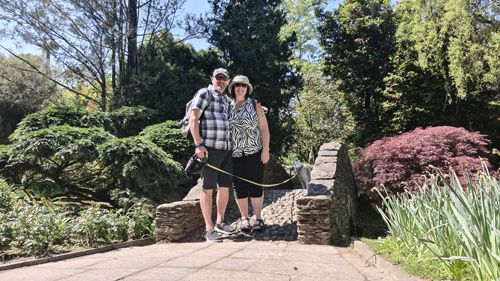 A man and woman are standing on a bridge in the Giardini Botanici di Villa Taranto, accompanied by their dog. The setting features outdoor elements like trees and plants, enhancing the natural beauty of the park. They are dressed appropriately for a stroll in a garden environment.