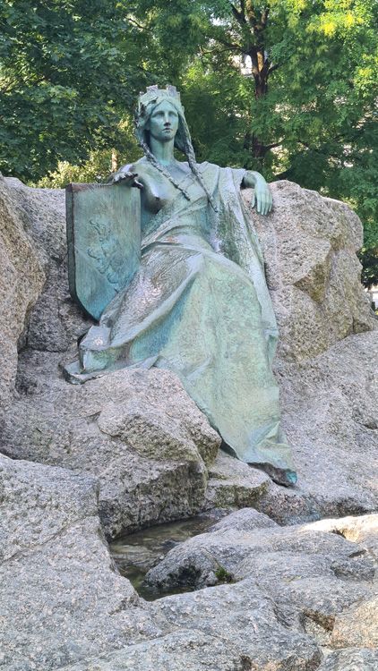 Statue of a person holding a book at the Universal Post Monument, surrounded by trees, buildings, and rock formations