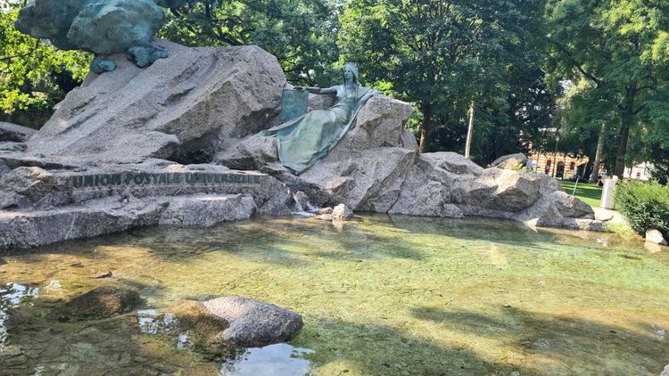 Large rock formation of the Universal Post Monument in Bern, set in a grassy area with trees, rocks, and nearby ponds