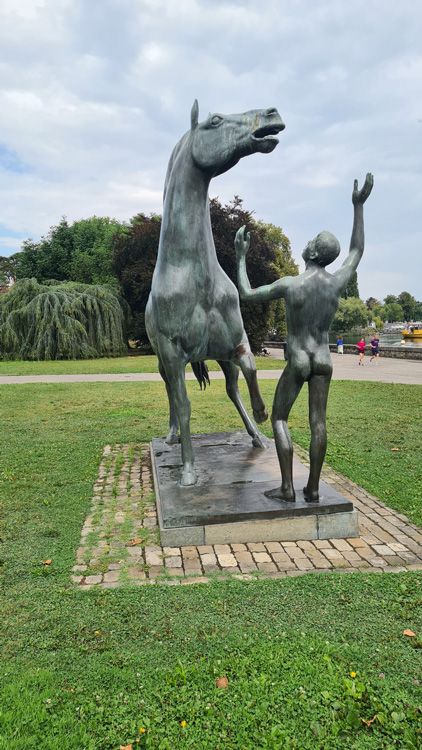 Bronze statue of two horses in a Geneva park, surrounded by grass, trees, and a cloudy sky, with a person standing beside it