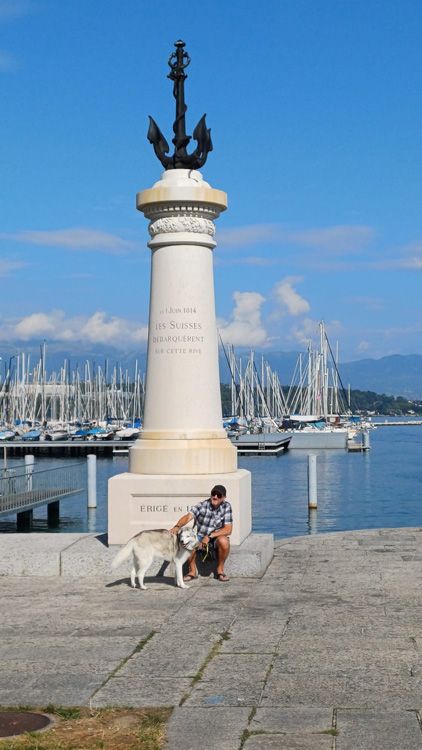 Person and dog seated on a bench in front of a statue at Port Noir, with lake and clear sky in the background