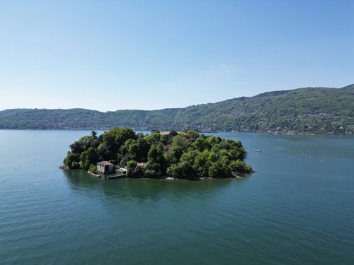 verdant island nestled in a serene lake. The island is densely covered with lush green trees and features a few small buildings. In the background, rolling hills extend to the horizon under a clear blue sky, suggesting a peaceful and possibly secluded location. A small boat is visible on the lake in the distance. The overall impression is one of tranquility and natural beauty. Lake Maggiore