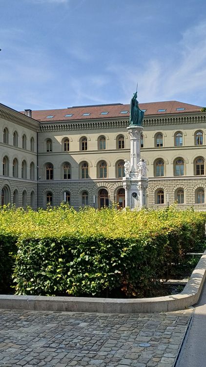 Large building with a prominent tower, stone facade, and surrounding garden area under a blue sky