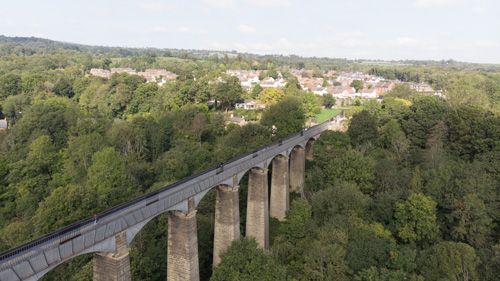 An aerial view of Pontcysyllte Aqueduct, a UNESCO World Heritage site in Wales, showcasing its iconic arched structure spanning the river Dee, with surrounding countryside and a clear sky in the background.