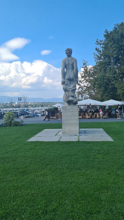 Statue in a park surrounded by open green space, trees, and clouds in the sky