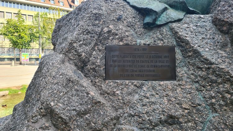 Plaque reading "PLU LES BETABITES BE CANTAR BE LA WILLE ET" affixed to a rock, surrounded by trees in an outdoor memorial area