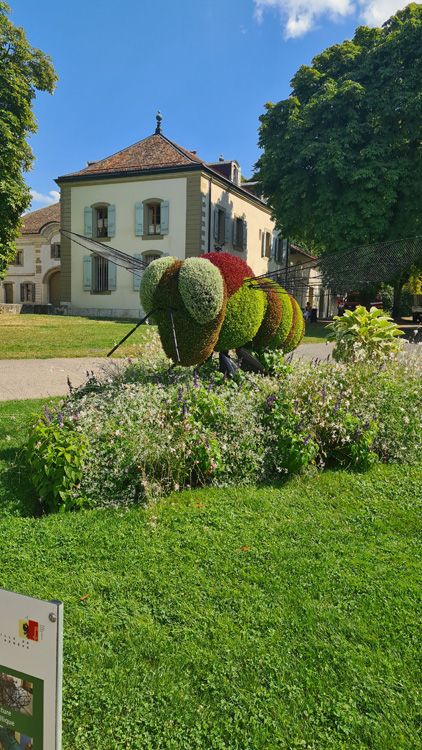 Large colorful bee sculpture in a vibrant garden with green grass, trees, shrubs, and a cloudy sky