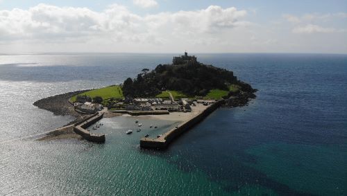 Experience the breathtaking aerial view of National Trust - St Michael's Mount. See the historic island castle, its gardens, and the surrounding Mount's Bay from a stunning bird’s-eye perspective.