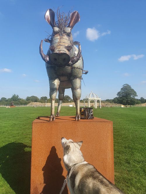 A dynamic sculpture at the British Ironwork Centre depicting a wild boar in a defensive stance, with a curious dog observing it closely