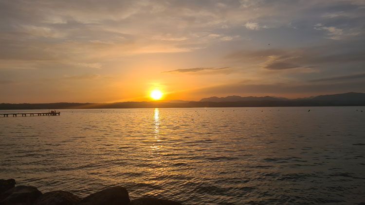 Peaceful sunset over a calm lake with hazy mountains, soft sky hues, a short pier, and subtle reflections on rippling water.