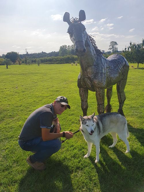 A whimsical metal sculpture of a house at the British Ironwork Centre, featuring figures of 'me' and 'Mia' admiring the artwork, set in the outdoor exhibition area.